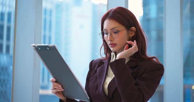 Close-up parallax shot of serious thoughtful Latina businesswoman adjusting glasses while checking project papers by office window with city view