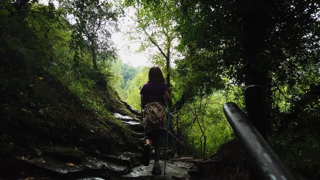 Young girl with long hair seen from behind hiking up a steep, wet stone path with a metal handrail, surrounded by a lush green forest with bright sunlight filtering through the trees