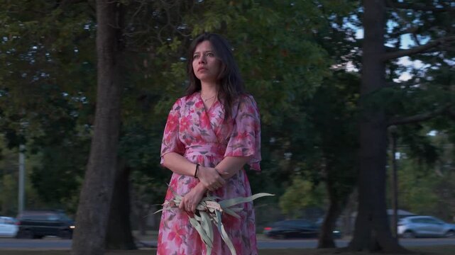 A woman in a vibrant floral dress stands peacefully in Croydon Park, enjoying the tranquil ambiance of the Australian landscape as evening shadows fall, captured in a smooth, steady shot.