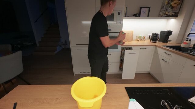 Person actively tidying up in a contemporary kitchen featuring clean lines and warm lighting. Focused on organizing with a bright yellow bin and sleek appliances. Medium shot.