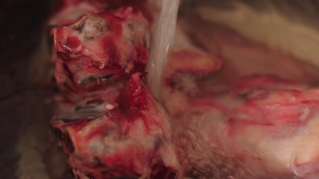 Macro shot: a stream of clean water pours onto fresh raw beef marrow bones with meat in a steel pot. Preparing natural ingredients for a rich meat broth or soup