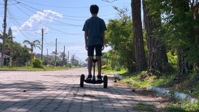 Child riding hoverboard down residential street on brick pavement, rear view with trees and power lines, low angle wide shot, moving away from camera, 4K30.