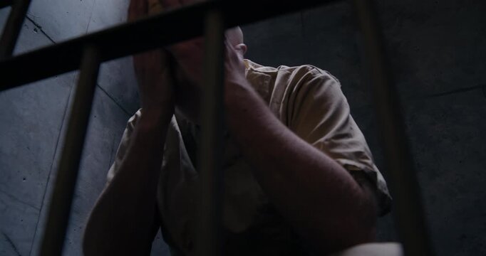 Low angle medium shot of prisoner with mask on in jail cell rubbing his head and face with his hands as the camera tracks right and pans left.