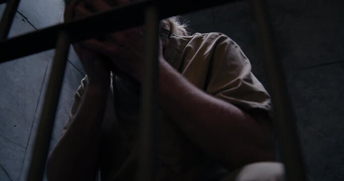 Low angle medium shot of prisoner with pony tail in jail cell rubbing face with hands as the camera tracks right and pans left.