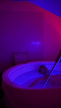 Young man floating in a saline sensory deprivation tank at Float Center Ljubljana in Ljubljana, Slovenia. Neon lighting, wellness spa therapy for deep relaxation and recovery, vertical timelapse