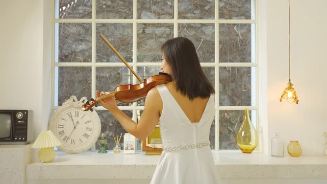 Back view talented woman playing the violin classic old style, violinist rehearsing her part before performance. Lady student with fiddle in light room by window, serene performance, classical music