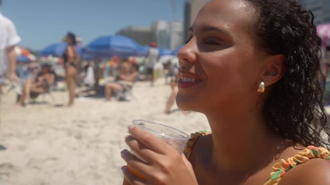 Young Latina woman sipping a yerba mate drink from a plastic cup and smiling, with blurred sun umbrellas and people in the background. Static close up clip at Copacabana beach, Brazil.