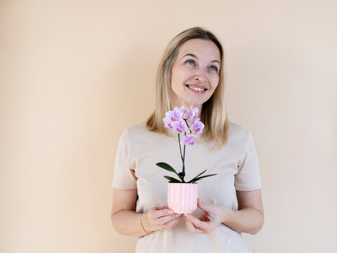 A blonde woman wearing a brown shirt is holding an orchid and standing in front of a brown background