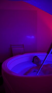 Man floats in a hyper saline float tank under blue and pink neon light, vertical static tripod shot.