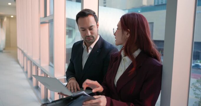 Professional Latina woman and European man in formal suits standing by office window and carefully analyzing paper documents with charts and data during sunset in city business center