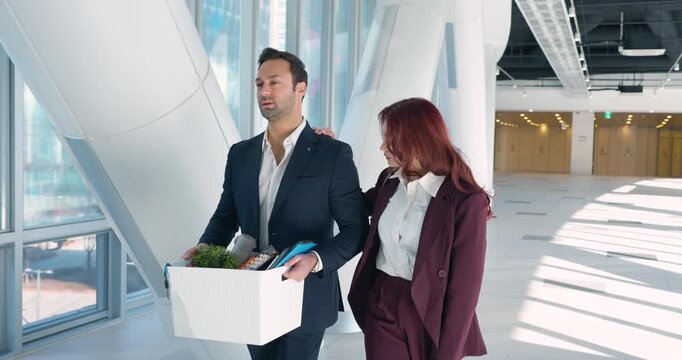 Sad European man walking with personal belongings in white box while Latina colleague comforts him in modern office corridor after termination or resignation