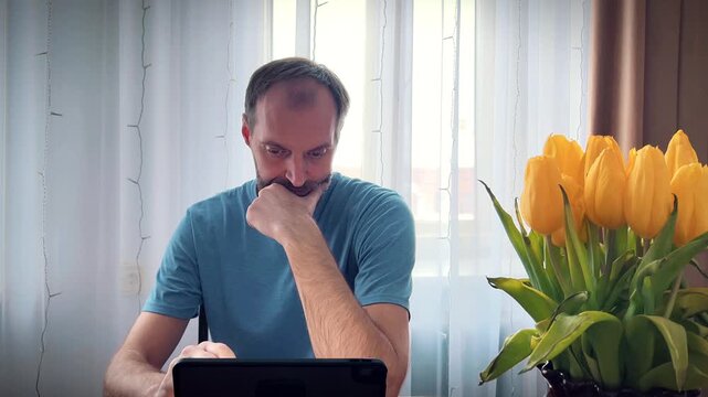 Mid-adult man in a blue t-shirt pondering over a digital tablet screen indoors. Versatile asset for remote work, problem-solving, and online education marketing materials.