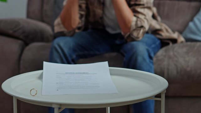Wife hand dropping wedding band falling onto legal divorce papers while distraught husband sits