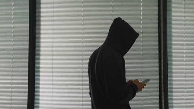 Hacker silhouette by window using phone to type message or secret email digital communication. Mysterious person with black hoodie indoor holding smartphone sending messages. Hacker or spy in a house
