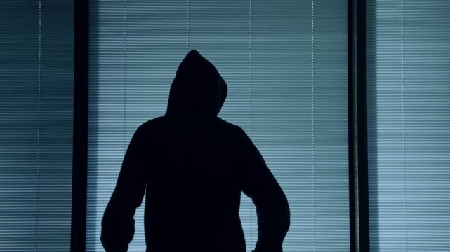 Silhouette of man with black hoodie standing by apartment window at night waiting or thinking of master plan. Hacker or spy mysterious person in suspicious clothes smart and pensive in back view