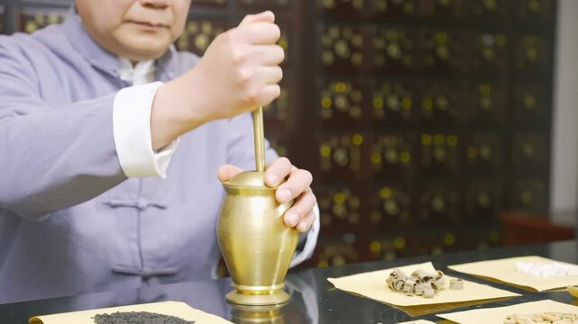 Herbalist apothecary oriental practitioner in traditional clothes doctor grinds dried herbs medicinal roots ingredients to make medicine in mortar, pestle for treatment surrounded by natural medicine