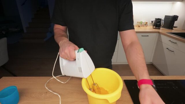 A man in a casual kitchen uses a hand mixer to blend ingredients in a bright yellow bowl, creating a warm and inviting atmosphere with soft overhead lighting.