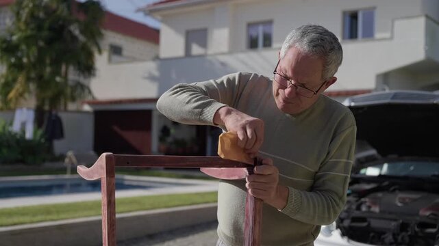 Elderly man wearing glasses sanding wooden chair frame outside near house and pool in daylight