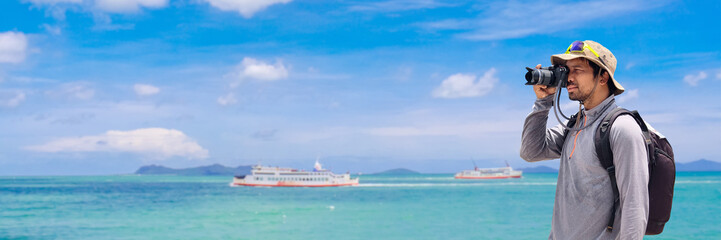 A man uses camera looking for the beautiful seascape view. He is wearing a backpack and hat.
