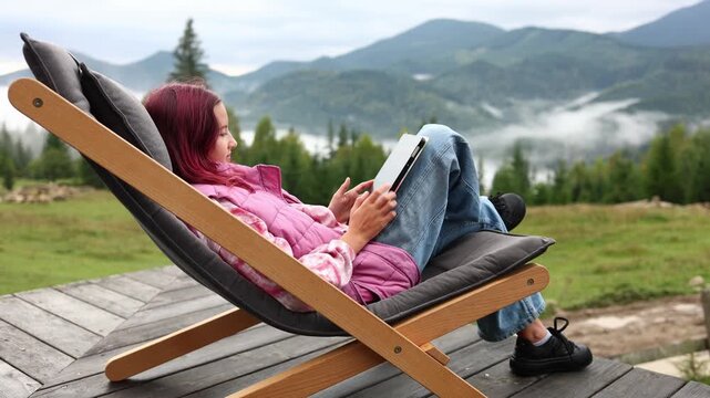 A teen girl sits comfortably on a deck chair with a tablet in her hands. She enjoys the mountain view surrounded by trees. It is a nice moment in her vacation.