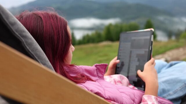 A teen girl sits comfortably on a deck chair with a tablet in her hands. She enjoys the mountain view surrounded by trees. It is a nice moment in her vacation.