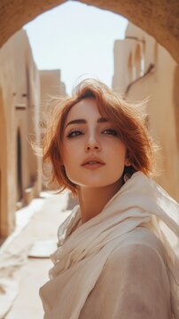 Selfie of an Iranian woman with short red hair and light makeup, wearing a white shawl, in the streets of old, adobe buildings