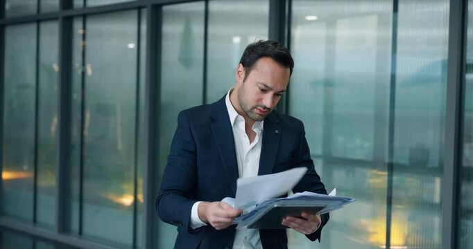 Stressed male professional in suit standing in glass office hallway looking frustrated while reviewing documents and bad financial data