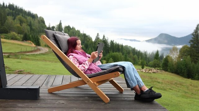 A teen girl sits comfortably on a deck chair with a tablet in her hands. She enjoys the mountain view surrounded by trees. It is a nice moment in her vacation.