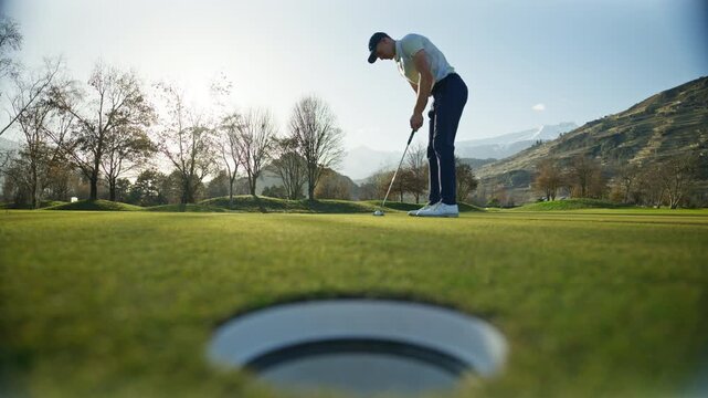 Closeup shot of a golf ball rolling across the green and dropping into the hole in Sion, Switzerland. Clean putting moment, precision sports action, and calm alpine golf course atmosphere.