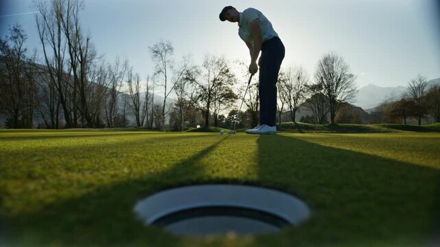 Wide shot of a golfer attempting a putt on the green in Sion, Switzerland, narrowly missing the hole. Calm golf course setting, scenic alpine surroundings, and authentic sports moment.