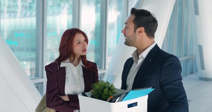 Dismissed male employee holding box of personal belongings stops to talk with female colleague standing arms crossed in bright modern corporate corridor