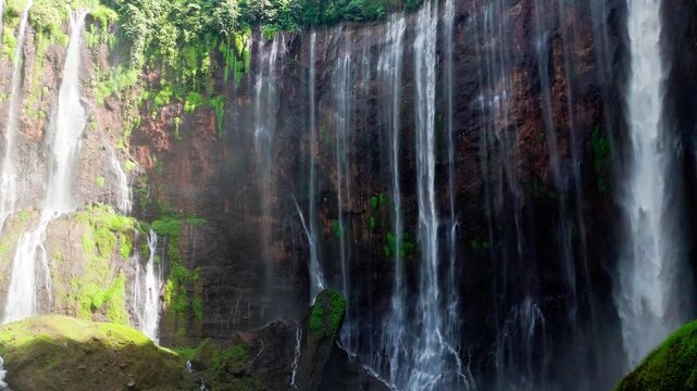 Epic 4K drone shot of Tumpak Sewu, Java, Indonesia. A couple walks toward the waterfall as the aerial camera rises, revealing towering canyon walls, flowing water, lush greenery, and misty natural bea