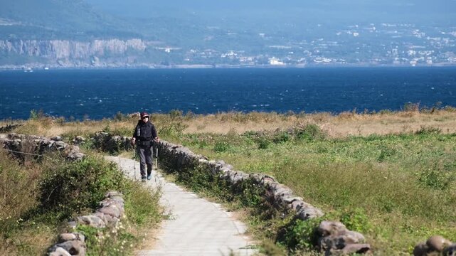 A hiker explores the captivating Olle Trail on Jeju Island. Surrounded by traditional stone walls and lush farmland, the path offers stunning ocean vistas under clear skies.