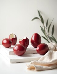 Fototapeta premium Glossy red Easter eggs rest on white geometric shapes. Some eggs are cracked revealing inner shells. Green leaves provide subtle natural accent to this festive food composition.