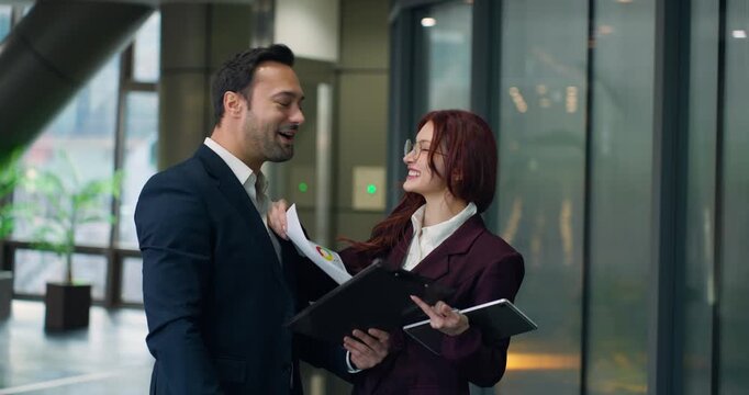 Two smiling business professionals share genuine laughter while holding documents and folders in bright modern corporate office corridor with glass walls
