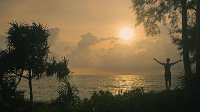 Silhouette of a person performing upward sun salute yoga pose at a tropical coastline under a tree during a golden sunset. Calm ocean and glowing sky creating a peaceful wellness scene.