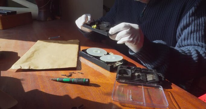 Specialist wearing white protective gloves carefully takes apart a VHS cassette shell using precision tools in a low-lit office environment.