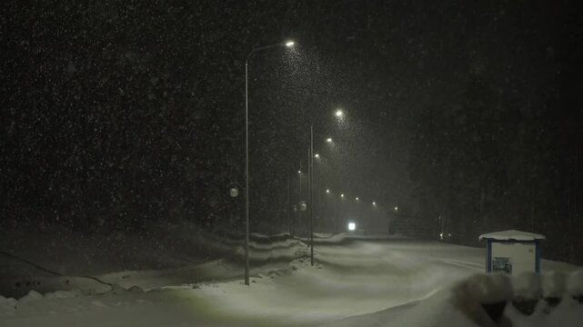 Deserted Snowy Road Under Lamplight Heavy Snowfall Blankets Asphalt, Snowdrifts Piling Along Roadside, Row Of Lampposts Casting Hazy Glow, Distant Headlights Piercing Flurries, Cold Cinematic Solitude