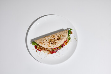 Fototapeta premium Tortilla wrap filled with vegetables, meat and cheese served on white plate on clean white background top view. Concept of street food, takeaway meal, casual dining and food delivery menu design.