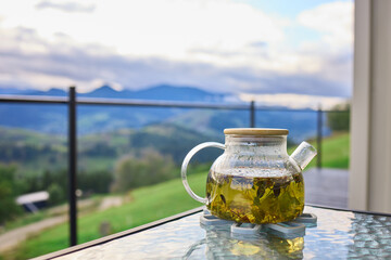Naklejka premium A glass teapot filled with herbal tea sits on a table. The background shows green mountains and a cloudy sky. It is a quiet setting for enjoying a warm drink.