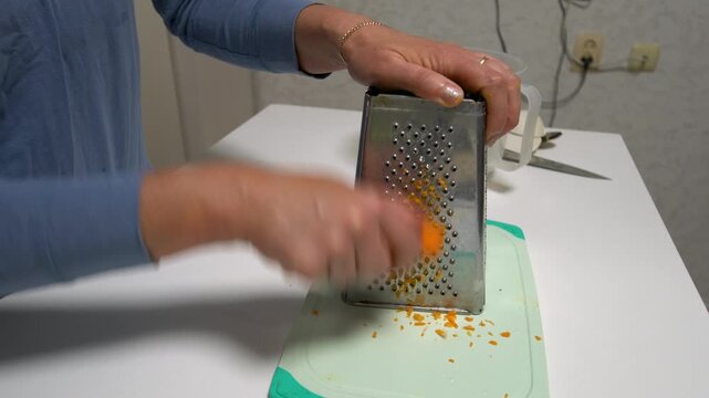 Hands are grating a carrot on a metal grater in a kitchen. A cutting board sits on the table, and tools are nearby.