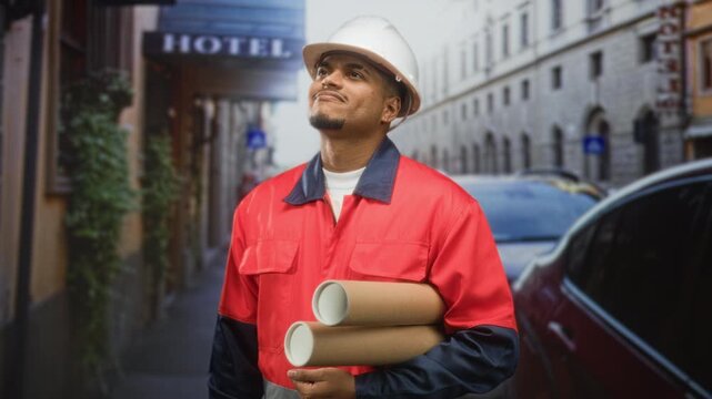 Hispanic man in hard hat holding rolled blueprints and wearing red uniform, beard visible, looks up on street near hotel; determination planning.