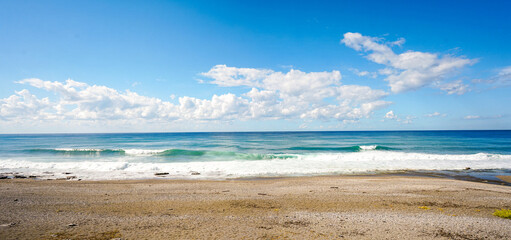 Wide-angle, tranquil view of a pebble beach and turquoise sea under a bright blue sky with white clouds. Clean coastal nature landscape offering ample copy space for design.