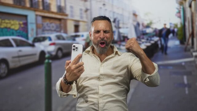 Man middle aged hispanic grey hair holding smartphone with clenched fist and shouting while pointing at the screen on a city street with parked cars and cafe tables; triumph celebration victory.