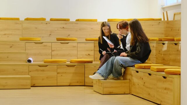 Teen study trio meets on wide wooden steps reviewing worksheet pages. Rolled paper rests on bench while teen group trades ideas writing answers