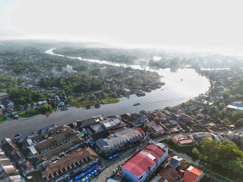 Aerial view of a vibrant riverside town with red-roofed houses, a main road, and a calm river under a clear blue sky