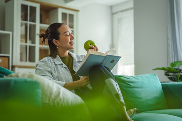 Young woman relaxing on couch reading book eating green apple