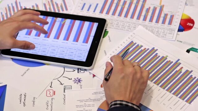Men's hands using a tablet and paper charts to analyze financial charts