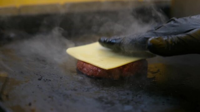 A close-up cinematic shot of a burger patty cooking on a grill with steam rising. A gloved hand carefully places a slice of cheese and then several pickle slices on top of the meat.