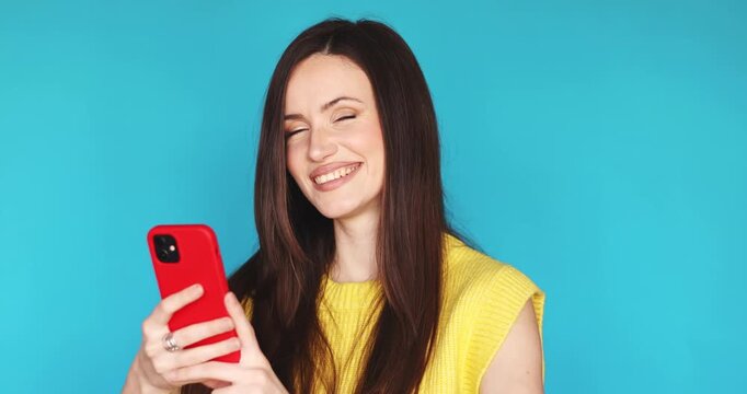 Proud young woman looking at red smartphone, smiling happily and pointing index finger at herself with confidence. Female achiever celebrating success or good news, isolated on blue background